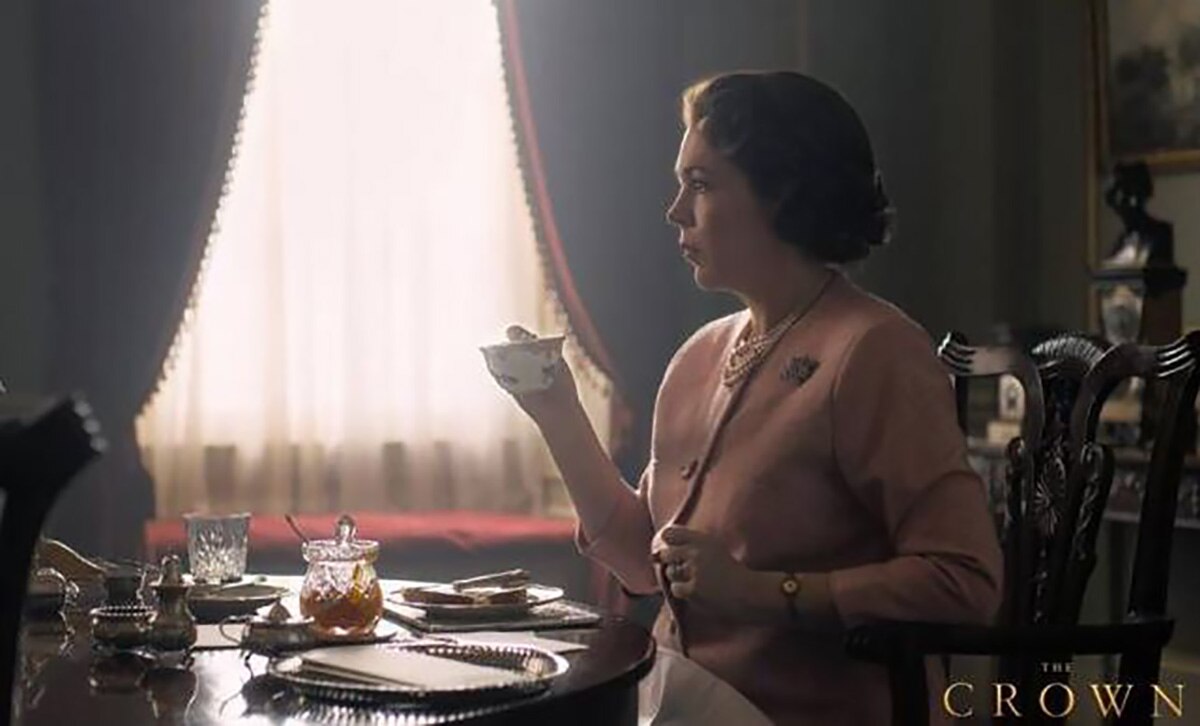Olivia Colman, que encarna a la reina Isabel, viste una chaqueta rosa, un collar de perlas y bebe una taza de té. FOTO: Especial
